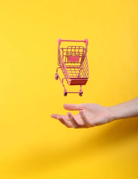 Hand Och Levitating Rosa Shoppingvagn Gul Bakgrund Minimalistiskt Stilleben Begreppskonst — Stockfoto