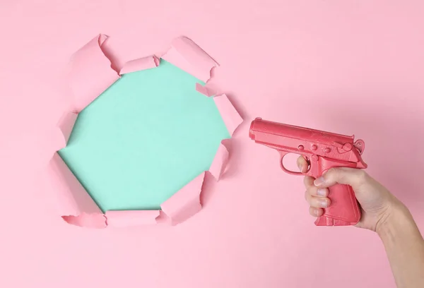 Hand holds pink pistol on blue-pink paper background with ripped space for copy space. Concept art, minimalism