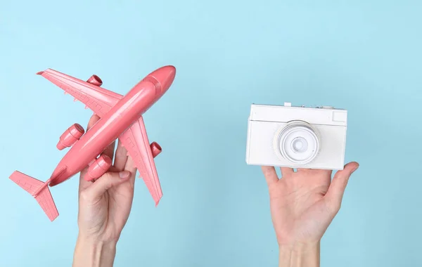 Mavi arka planda beyaz kamera ve pembe uçak tutan eller. Minimalizm seyahat kavramı