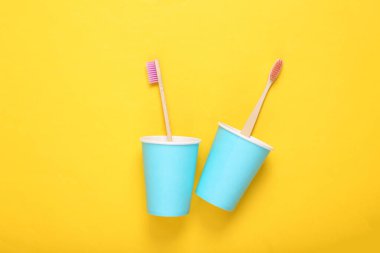 Sarı arka planda karton bardaklarda iki eko bambu diş fırçası.