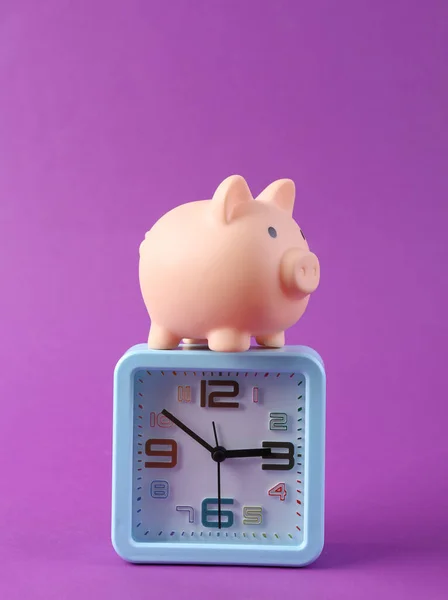 Alarm clock with piggy bank on purple background