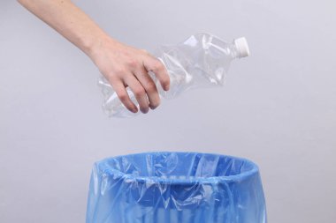 Elleri buruşmuş plastik şişeyi gri arka planda bir paketle çöp kutusuna atıyor.