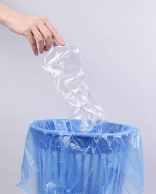 Elleri buruşmuş plastik şişeyi gri arka planda bir paketle çöp kutusuna atıyor.