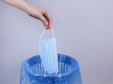 El, tıbbi maskeyi gri arka planda paketli çöp kutusuna atar.