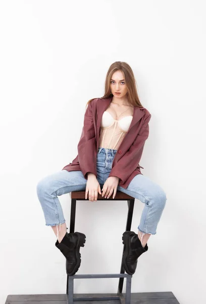 时尚女模特 穿着牛仔裤 皮夹克和紧身衣坐在椅子上 白色背景 — 图库照片