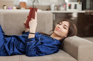 Gecelikli genç kadın oturma odasındaki kanepede uzanmış kitap okuyor.