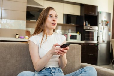 Keskin bir kadın televizyon ekranına bakar ve televizyon kanallarını uzaktan kumandayla değiştirir. Oturma odasındaki bir kanepede oturur..