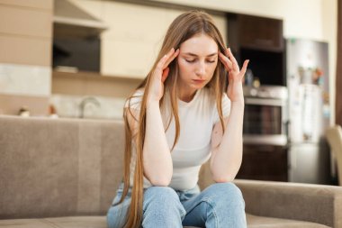 Baş ağrısı. Genç bir kadın oturma odasında kanepede otururken başını tutuyor.
