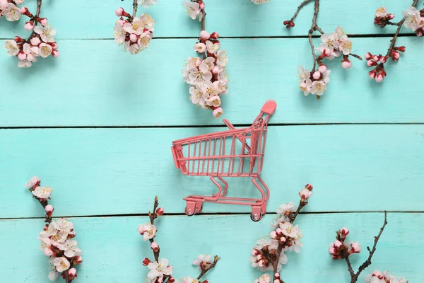 Mini alışveriş arabası ve mavi ahşap arka planda güzel pembe çiçekli dallar. İlkbahar konsepti. Düz yatış, üst görünüm