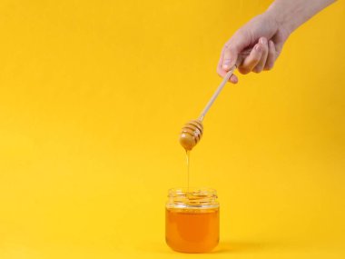Sarı arka planda bal kavanozuyla bal kaşığı tutan eller.