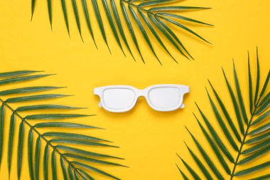 Sarı arka planda beyaz güneş gözlüğü ve palmiye yaprakları. Tropikal, asgari yerleşim. Üst Manzara. Düz yatıyordu