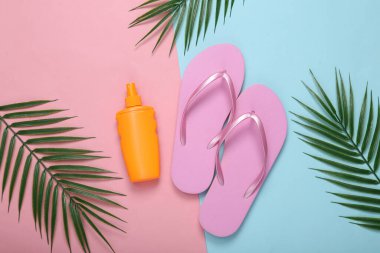 Güneş kremi şişesi ve parmak arası terlikler, pembe mavi arka planda palmiye yaprakları. Tropik, yaz uykusu kompozisyonu. Üst Manzara. Düz yatıyordu