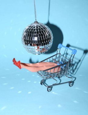 Bebek bacakları mavi parlak arka planda disko topunun altında alışveriş arabasında. Minimalizm partisi konsepti