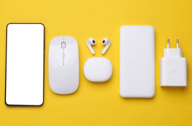 Modern aletler. Kablosuz kulaklıklar, şarj aleti adaptörü, bilgisayar faresi, akıllı telefon ve sarı arka planda elektrik bankası. Düz yatıyordu