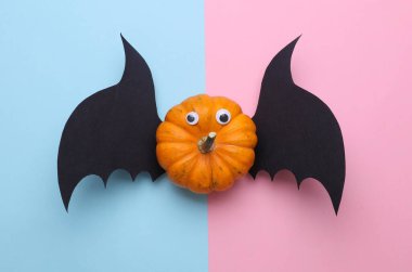 Pembe mavi arka planda, şaşı gözlü ve yarasa kanatlı balkabağı. El yapımı Cadılar Bayramı dekoru