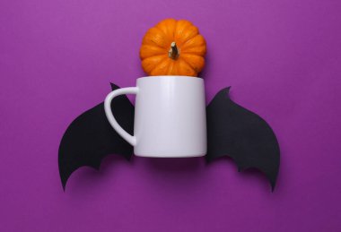 Tasarımınız için beyaz seramik kupa ve mor arka planda Cadılar Bayramı dekoratif yarasa kanatları ve balkabağı.