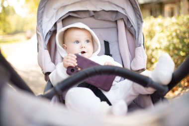 Kapüşonlu küçük şirin bebek akıllı telefonuyla oynuyor ve dışarıda bebek arabasında oturuyor.