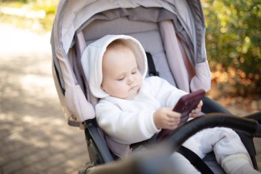 Kapüşonlu küçük şirin bebek akıllı telefonuyla oynuyor ve dışarıda bebek arabasında oturuyor.