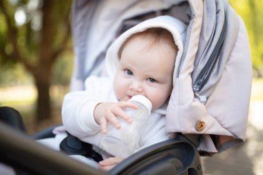 Kapüşonlu bir bebek şişeden su içiyor ve bebek arabasında oturuyor.
