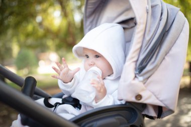 Kapüşonlu bir bebek şişeden su içiyor ve bebek arabasında oturuyor.