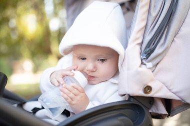 Kapüşonlu bir bebek şişeden su içiyor ve bebek arabasında oturuyor.