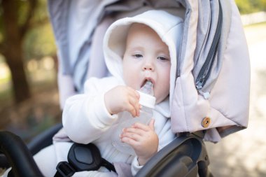 Kapüşonlu bir bebek şişeden su içiyor ve bebek arabasında oturuyor.