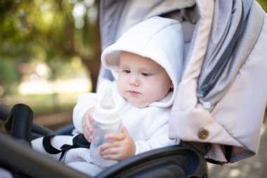 Kapüşonlu bir bebek şişeden su içiyor ve bebek arabasında oturuyor.