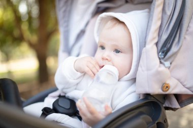 Kapüşonlu bir bebek şişeden su içiyor ve bebek arabasında oturuyor.