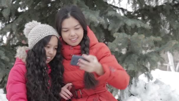 Sonriendo a dos chicas tomando una selfie con teléfono inteligente al aire libre en ropa de abrigo — Vídeo de stock