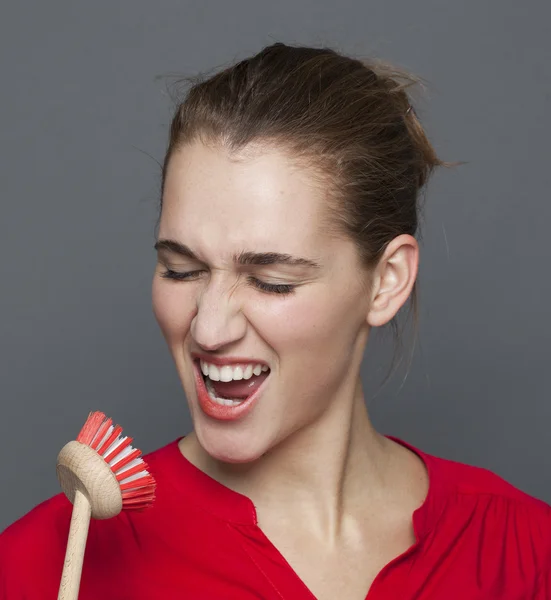 Excited young cleaning lady singing on a dish brush microphone — Stock Photo, Image