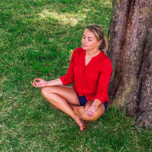 Bronzeada menina loira meditando sob uma árvore na grama — Fotografia de Stock