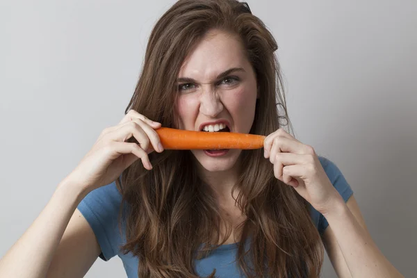 Hungry 20s girl biting a carrot with appetite — Stockfoto