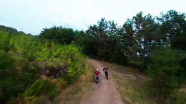 Exciting FPV drone chase of two motocross riders competing on a forest dirt track. Riders jump ramps, race at high speed, and showcase extreme motocross action from a cinematic aerial perspective.