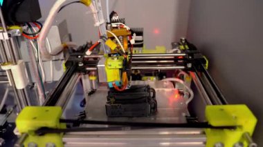 Bir 3D yazıcı erimiş filament üretim teknolojisini kullanarak plastik parçaları basar. Parmak izi başlığı, erimiş maddeleri cam bir levhanın üzerine bırakarak ayrıntılı siyah bileşenler oluşturuyor. Sahnede modern mühendislik, otomatikleştirilmiş imalat sergileniyor
