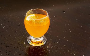 A close-up of a round glass filled with a vibrant orange drink. Garnished with finely ground spices and ice cubes on wet surface. Selective focus.