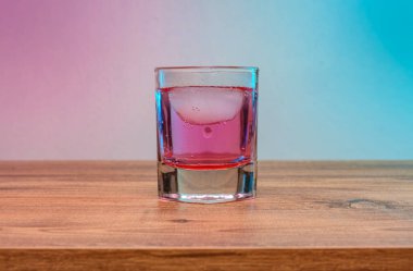 Close-up of a single glass filled with a vibrant pink cocktail. Featuring ice cubes and a fizzy texture. Set against a colorful gradient background of pink and blue hues.