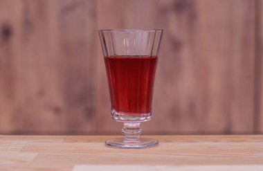 Red drink in an elegant glass goblet. Glass filled with a deep red beverage stands gracefully on a wooden table against a blurred wooden background. Selectice focus.
