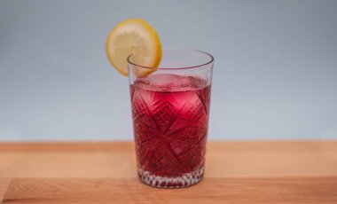 Refreshing iced red beverage with lemon slice on wooden table. An intricately patterned glass. Summer beverages, recipes and lifestyle concepts. Selective focus.