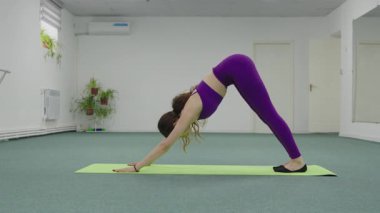 Genç bir kadın vinyasa sekansından aşağı bakan bir köpekten, spor salonundaki yeşil bir paspasın üzerinde kobra pozuna geçer..