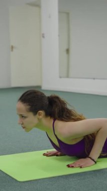 Side view of a young woman doing upward facing dog or cobra stretch on a green yoga mat in a bright studio.