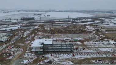 Gri gökyüzünün altında yeni bir yapı iskeleti ve karlı tarlaları olan bir inşaat alanının geniş hava manzarası..