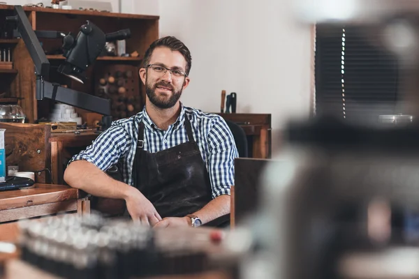 Jeweler in apron sitting at bench full of tools — Stock Photo, Image