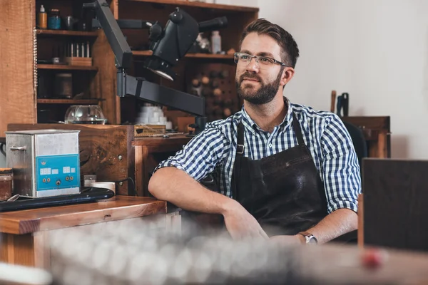 Jeweler in apron sitting at bench full of tools — Stock Photo, Image