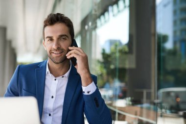Gülümseyen genç işadamı bir kafede otururken cep telefonuyla konuşuyor ve dizüstü bilgisayarla çalışıyor.