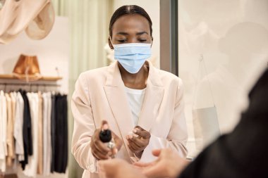 Koruyucu maskeli Afrikalı Amerikalı kadın girişimci dükkanının girişindeki müşterinin ellerine dezenfektan sürüyor.