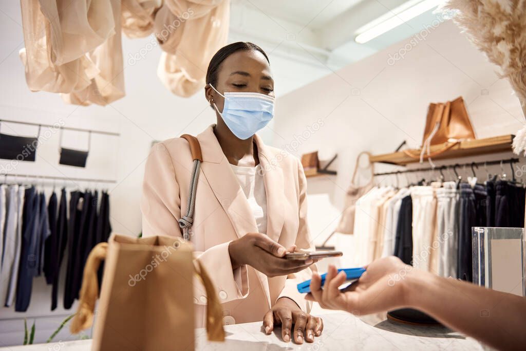 African American female customer in a protective face mask making a contactless payment using her smart phone in a clothing boutique