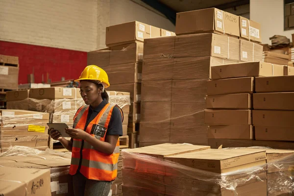 Young African female warehouse worker with a digital tablet doing an inventory check of boxes on pallets