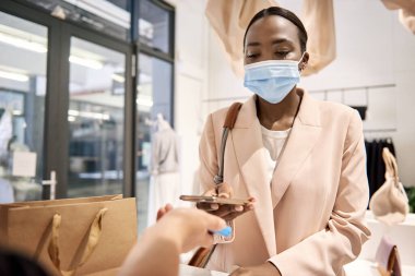 Koruyucu maskeli Afro-Amerikan bayan müşteri akıllı telefonunu bir butikte ulaşılamaz ödeme için kullanıyor.