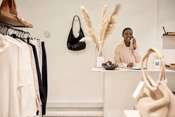 年轻的非洲裔美国女企业家笑着在时尚服装店的柜台边打电话 — 图库照片