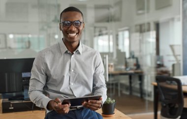 Ofiste masasına yaslanıp tablet üzerinde çalışırken gülümseyen genç bir Afrikalı işadamının portresi.